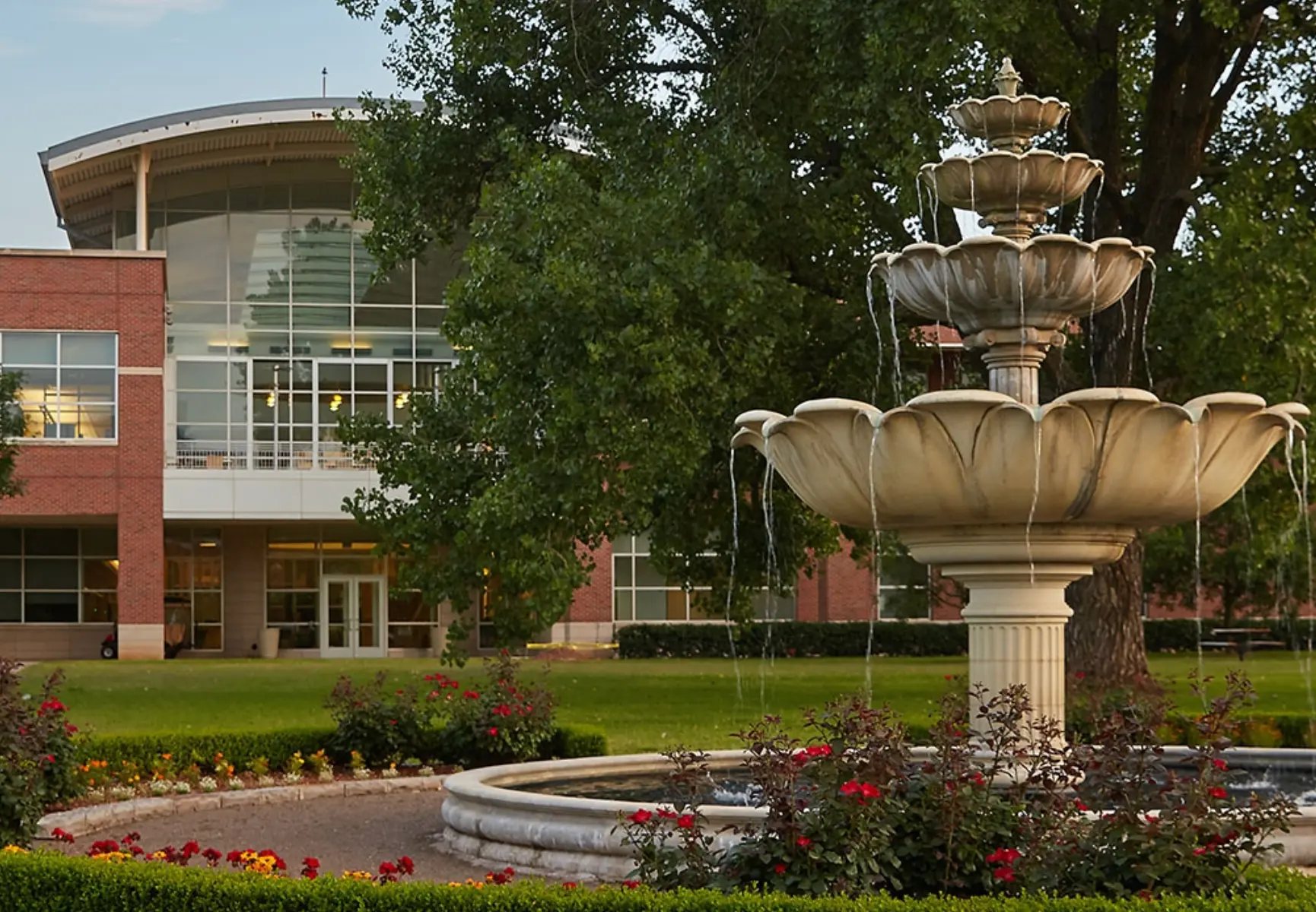 A four-tiered stone fountain standing in a landscaped garden of red roses and a low clipped hedge, with a two-story red-brick building behind it whose curved wall of glass windows reflects a tall light-colored tower, mature green trees, a manicured lawn, and a pale hazy sky
