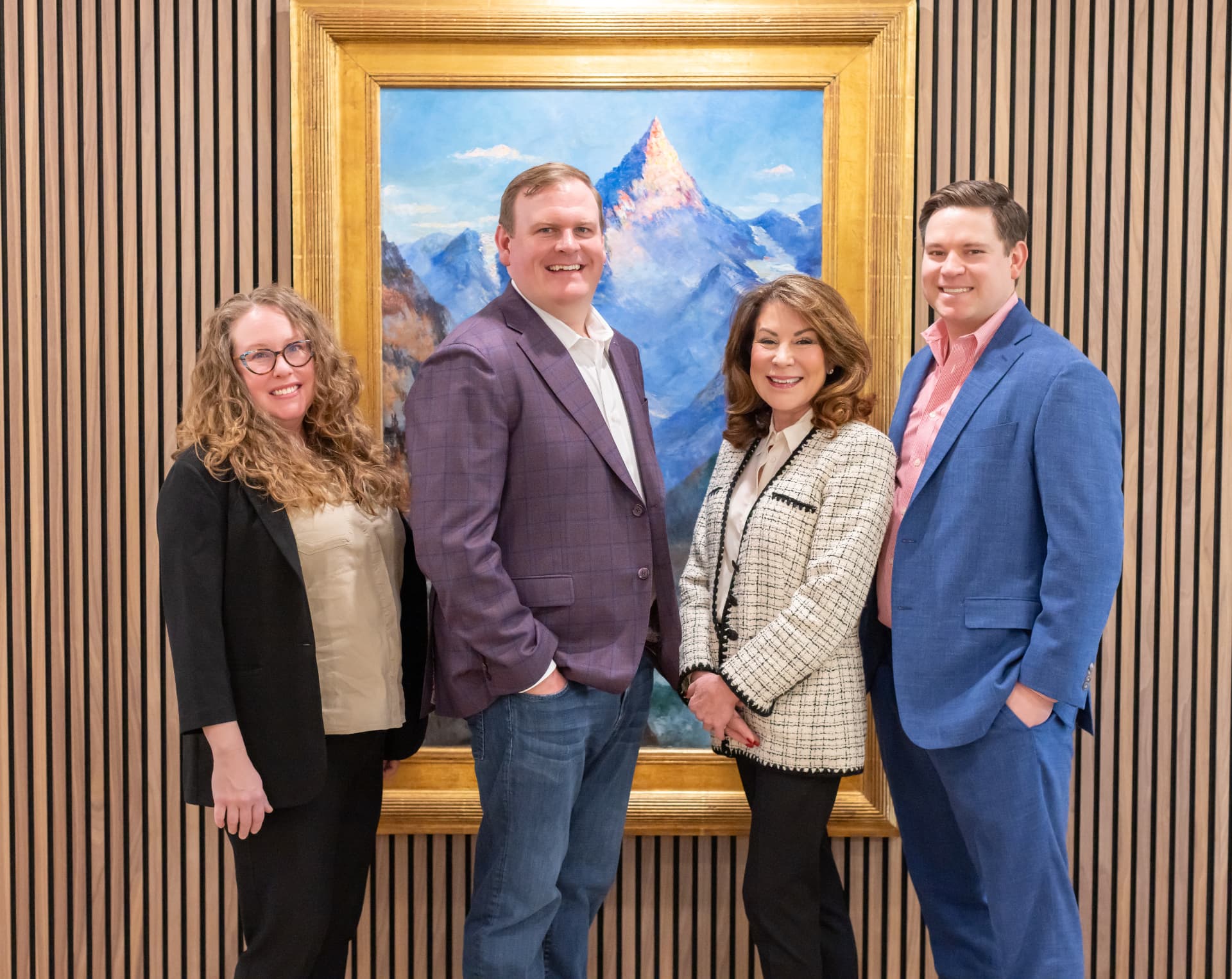Four people standing side by side and smiling in front of a large gilded-framed painting of a snow-capped mountain, hung on a wall of warm wooden slats