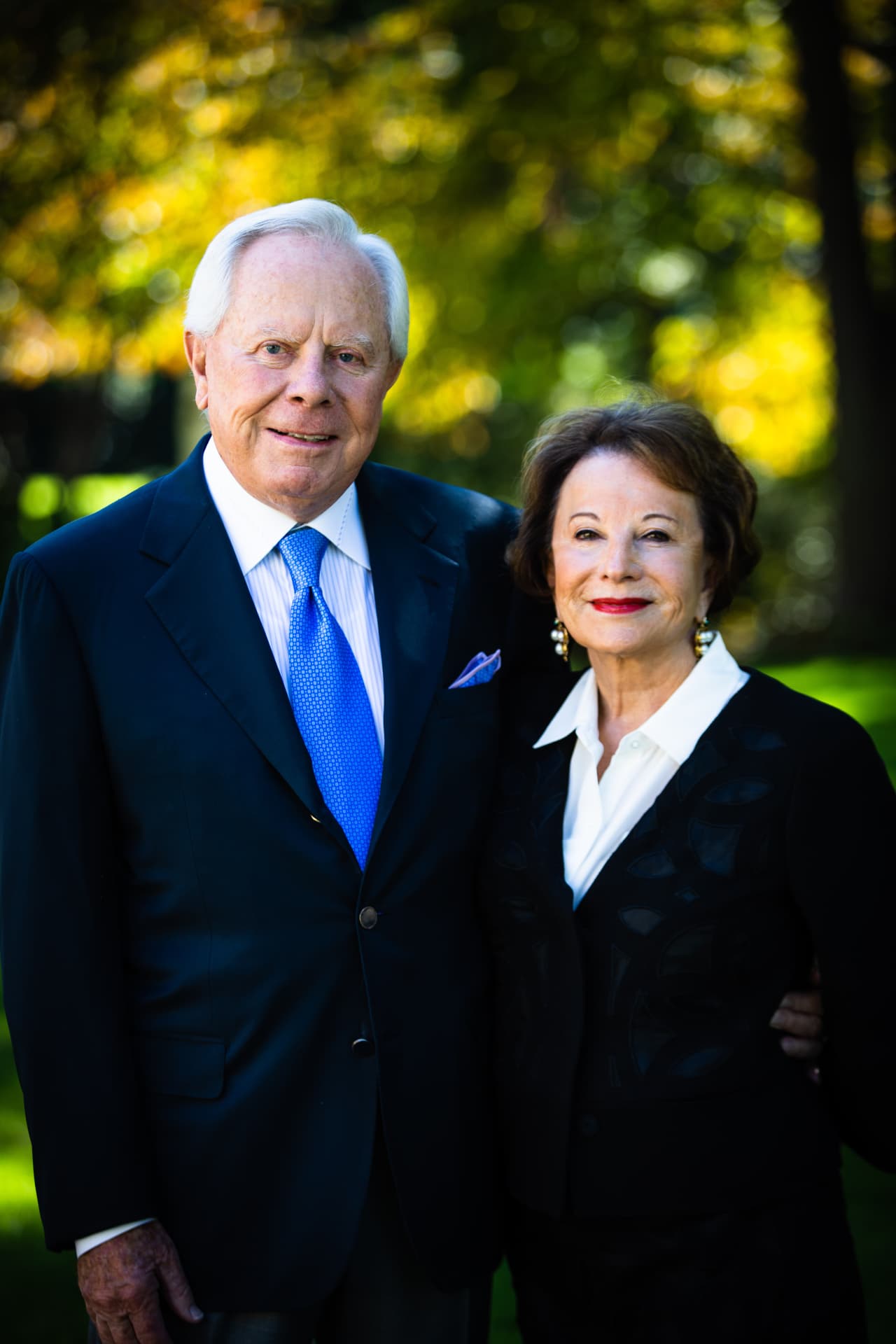 Peggy and Charles Stephenson standing close together outdoors in front of sunlit trees; Charles wears a dark suit with a blue tie and a lavender pocket square, and Peggy a black jacket over a white blouse