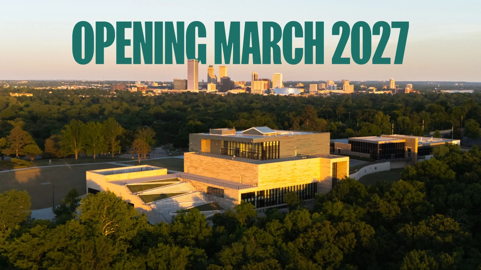 Aerial view of the new Gilcrease Museum building in warm golden light, surrounded by mature trees, with the downtown Tulsa skyline on the horizon and the words “Opening March 2027” across the sky