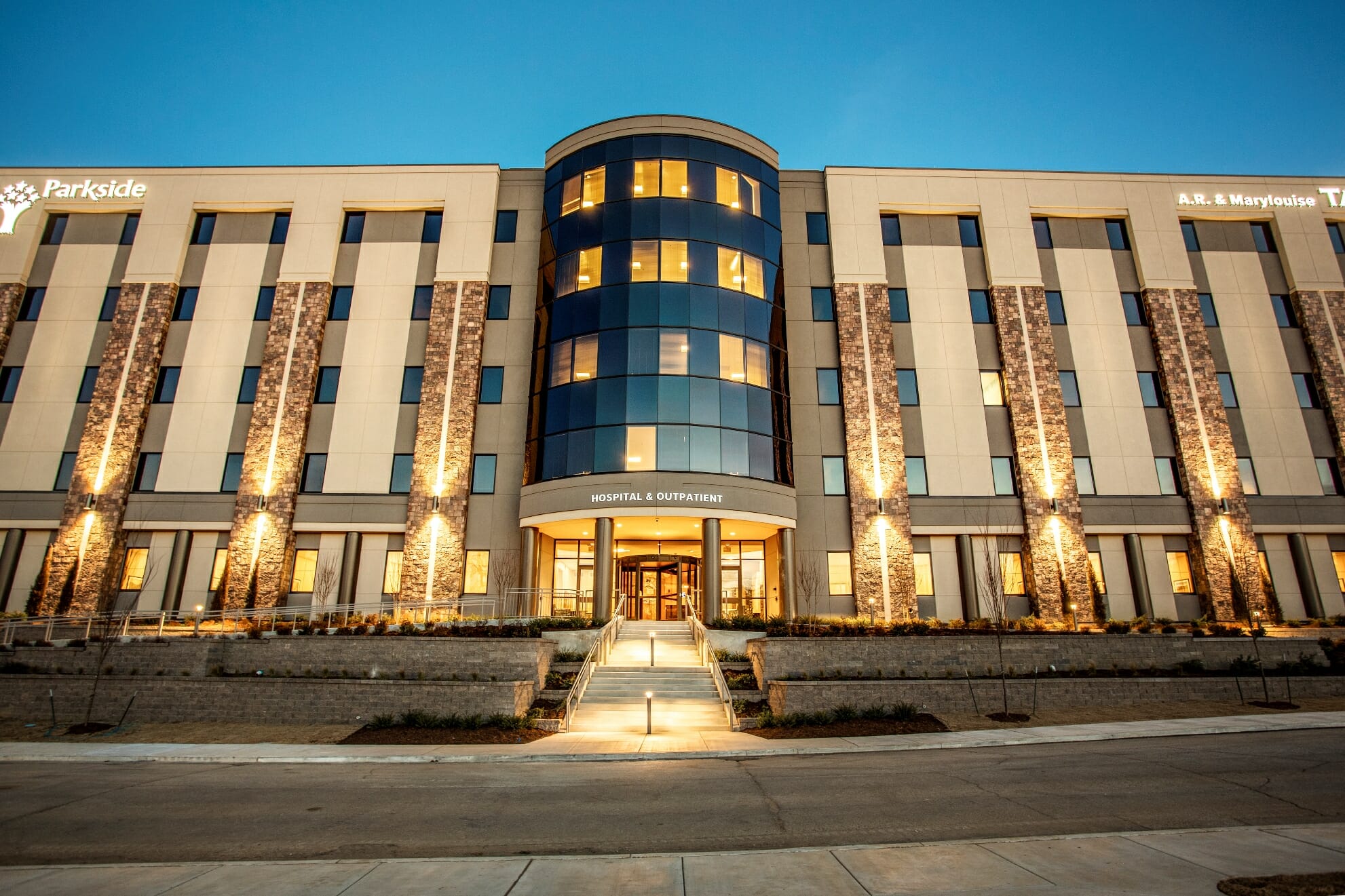 The Parkside hospital at dusk — a multi-story building with a curved glass entrance tower glowing with warm interior light, off-white wings faced with brown stacked-stone piers, a concrete staircase with metal handrails and small bollard lights leading up to the entrance canopy, and the words “Parkside” and “HOSPITAL & OUTPATIENT” on the building against a clear blue twilight sky