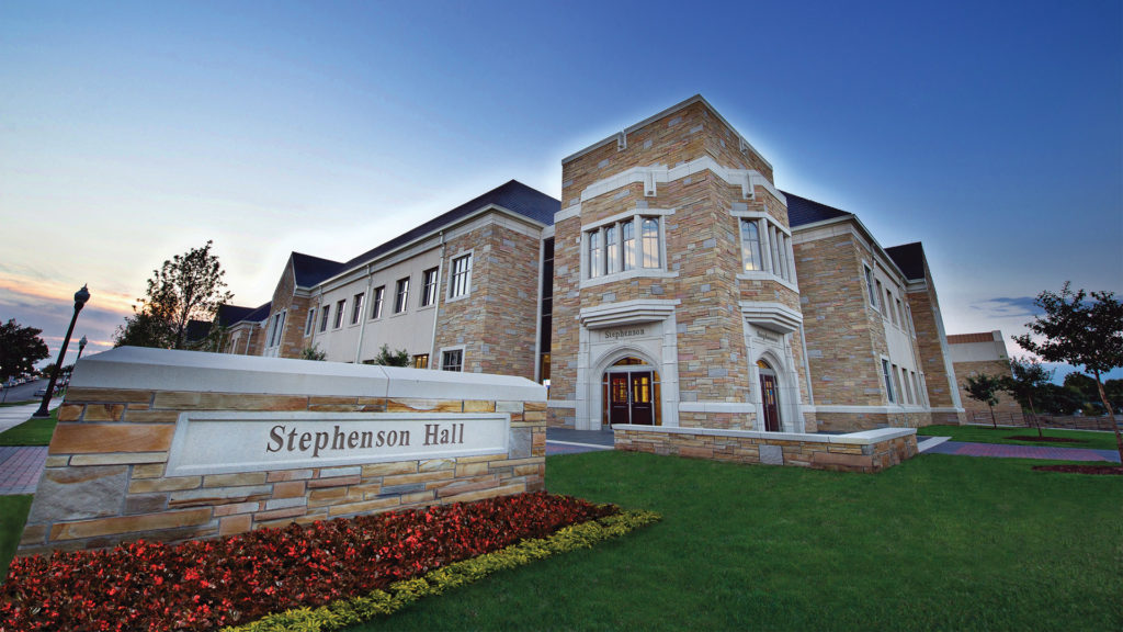Stephenson Hall at dusk — a stone academic building with a faceted entrance tower whose limestone arch is carved with “Stephenson”, a monument sign reading “Stephenson Hall” set in a bed of red flowers, a green lawn in front, and a blue sky fading to peach at the horizon