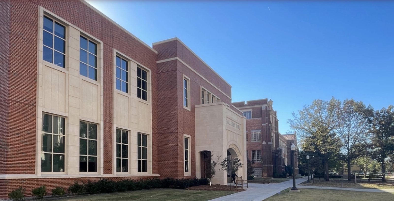 Gallogly Hall on the University of Oklahoma’s Norman campus — a red-brick building with cream limestone trim, an arched stone entrance with “GALLOGLY HALL” carved above it, a lamppost, a bench, and a walkway lined with mature trees under a clear blue sky
